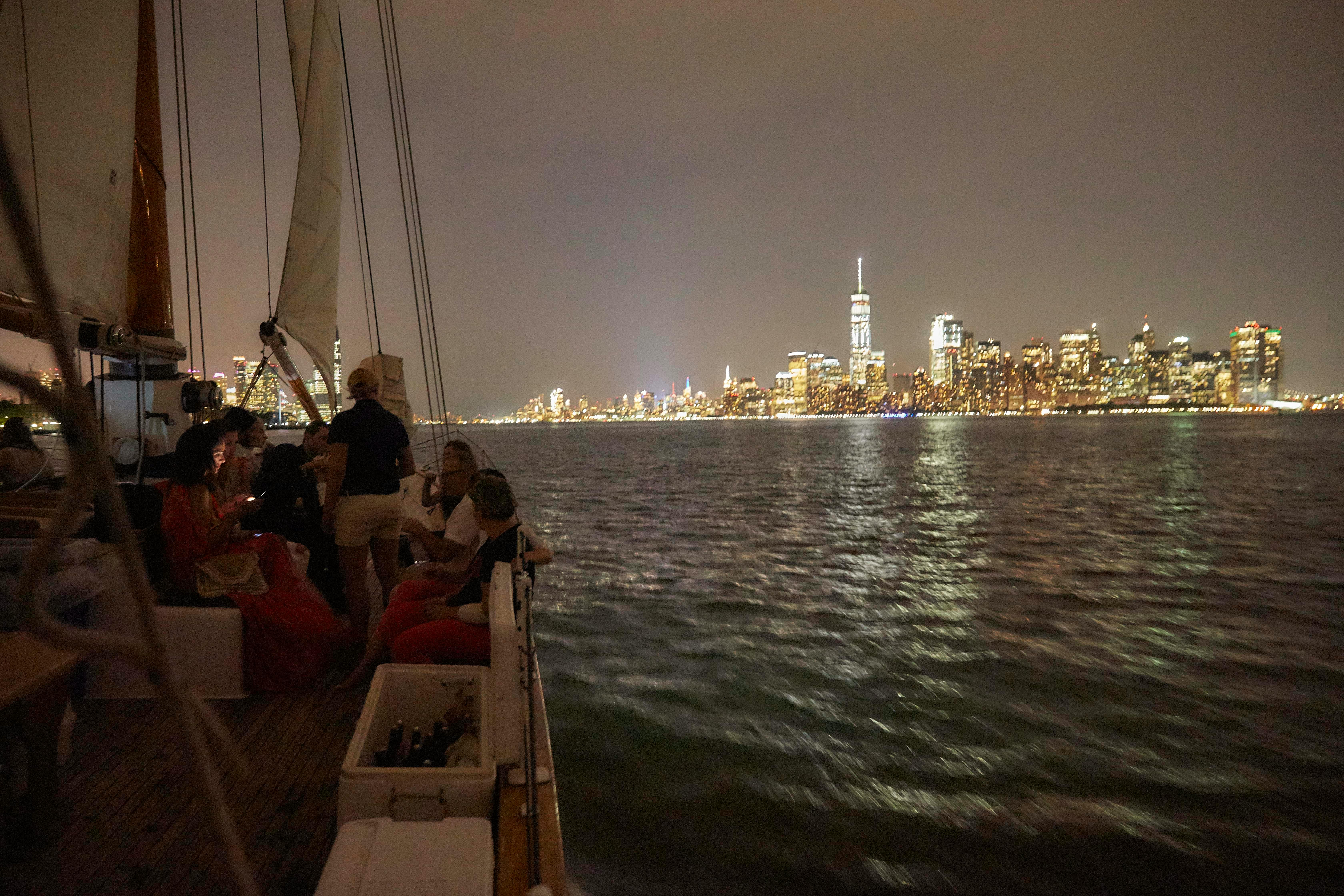 Άνθρωποι σε ένα ιστιοφόρο σε ένα λιμάνι τη νύχτα με τον φωτισμένο ορίζοντα της πόλης στο παρασκήνιο.