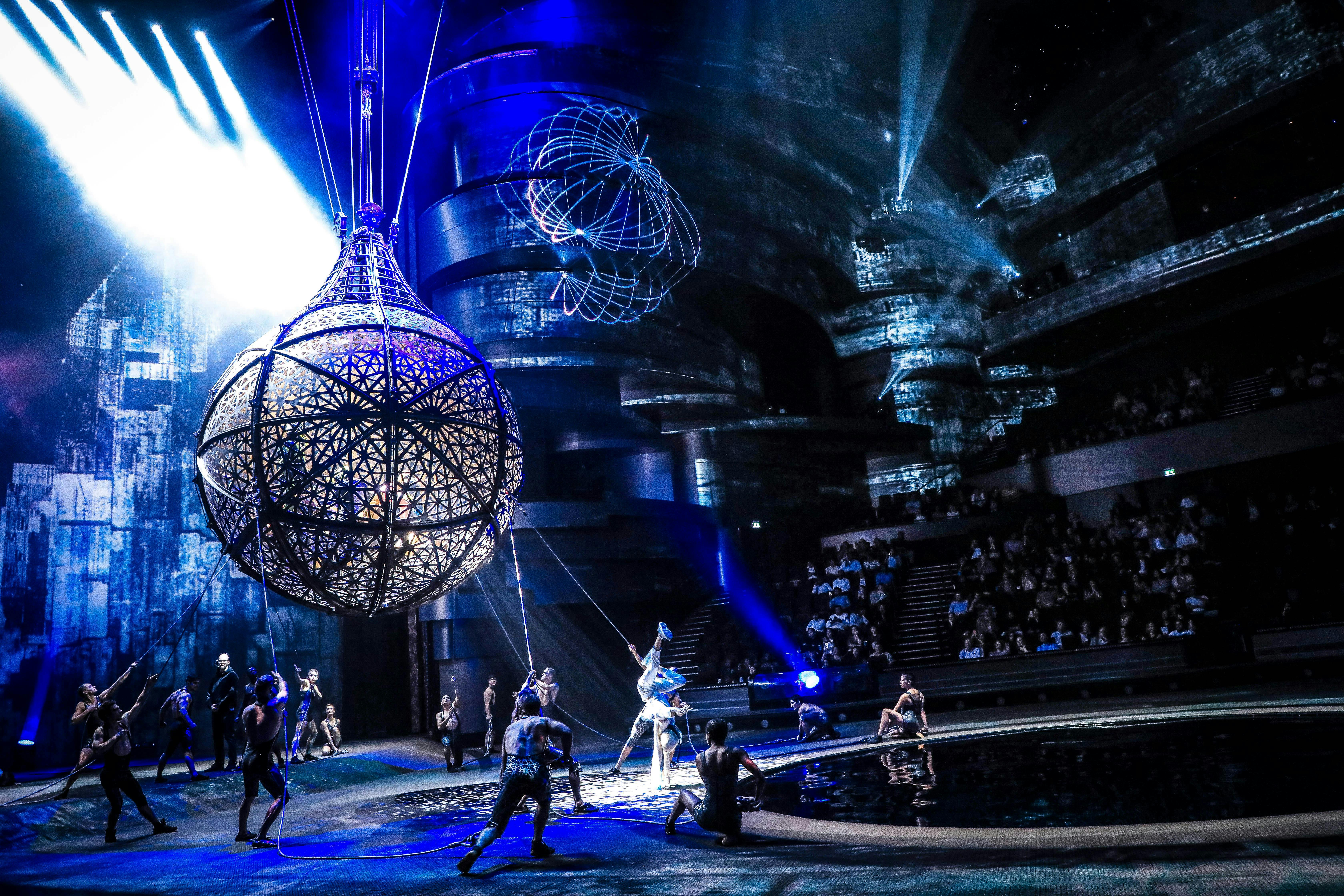 Gli acrobati si esibiscono intorno a una grande struttura sferica illuminata su un palcoscenico poco illuminato con una tonalità blu, di fronte a un pubblico.