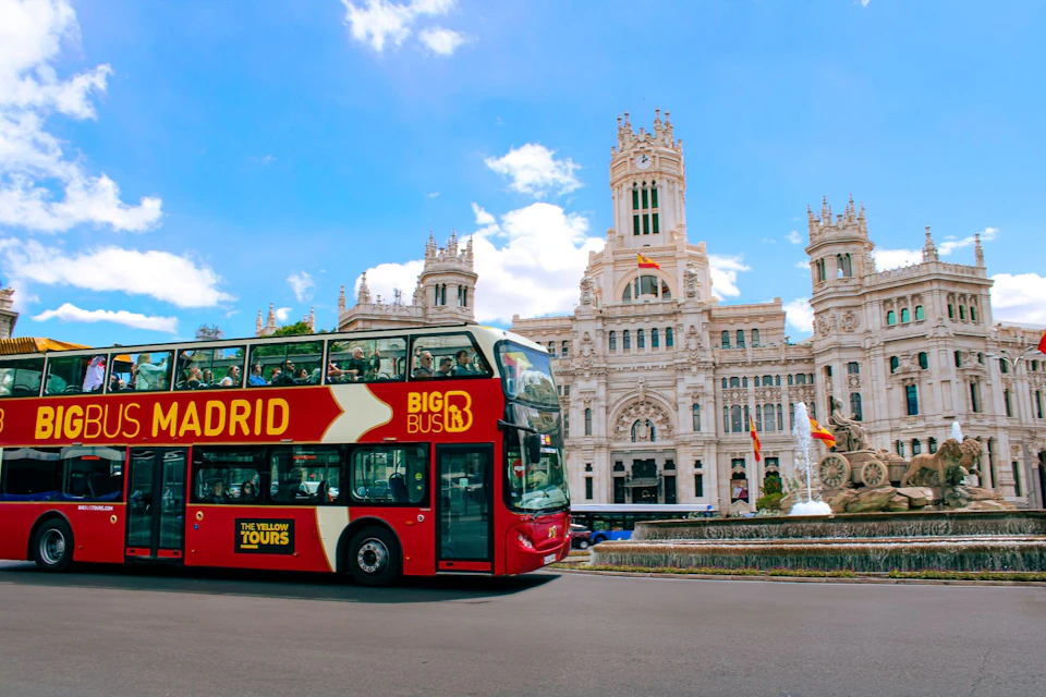 A red double-decker tour bus labeled "Madrid" passes in front of an ornate white building under a clear blue sky.
