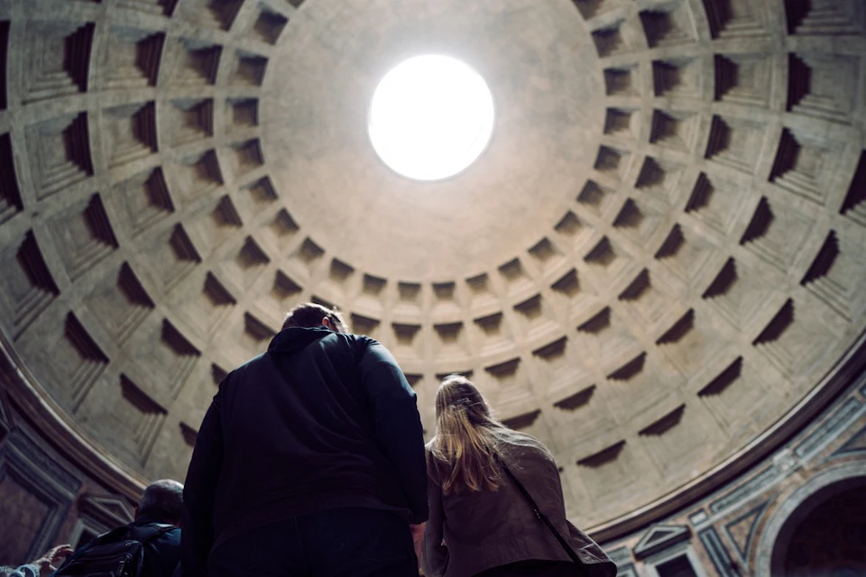Two people look up at a large, circular, dome ceiling with a central oculus, inside a grand architectural structure.