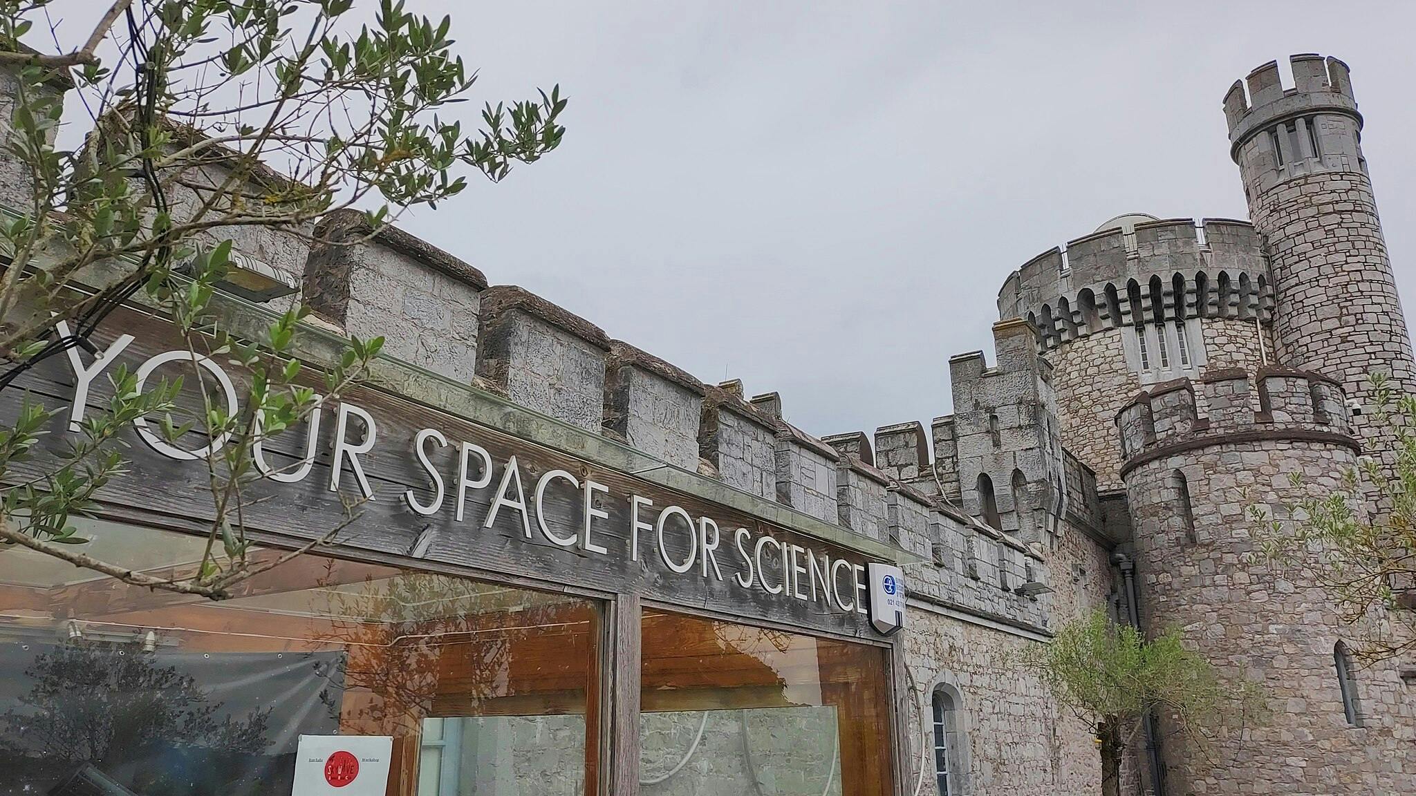 塔のある石造りの城と、「科学のためのあなたのスペース」と書かれた看板が付いたモダンな建物。 前景にはまばらな緑。