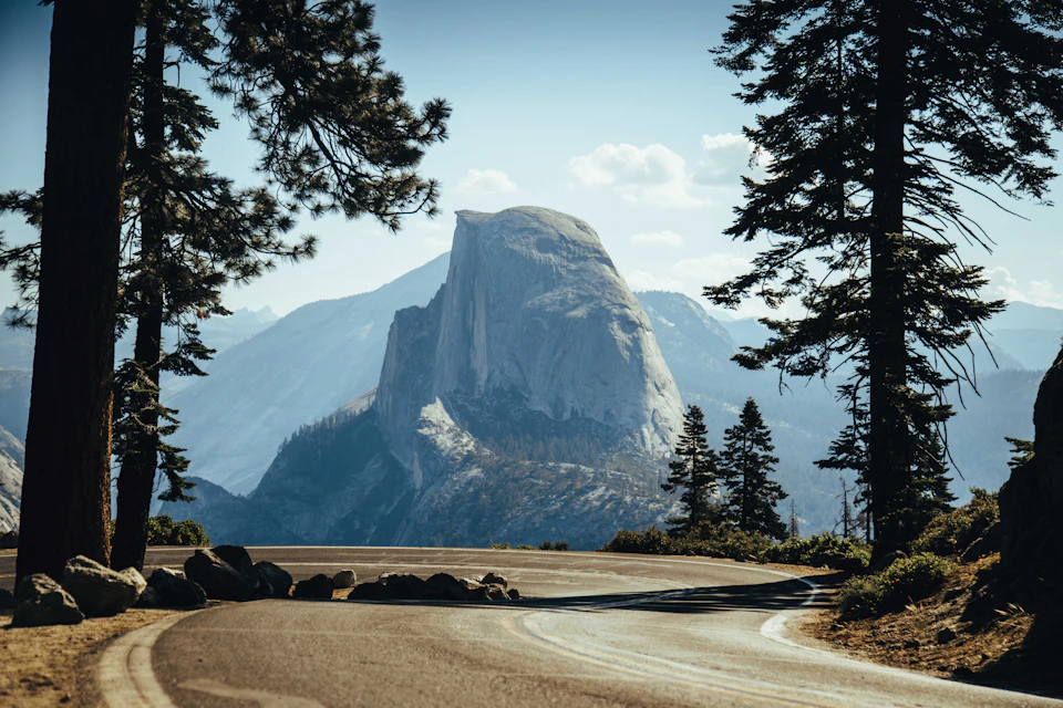 Curved mountain road encircled by tall pine trees, with a large, dome-shaped rock formation in the background under a clear sky.