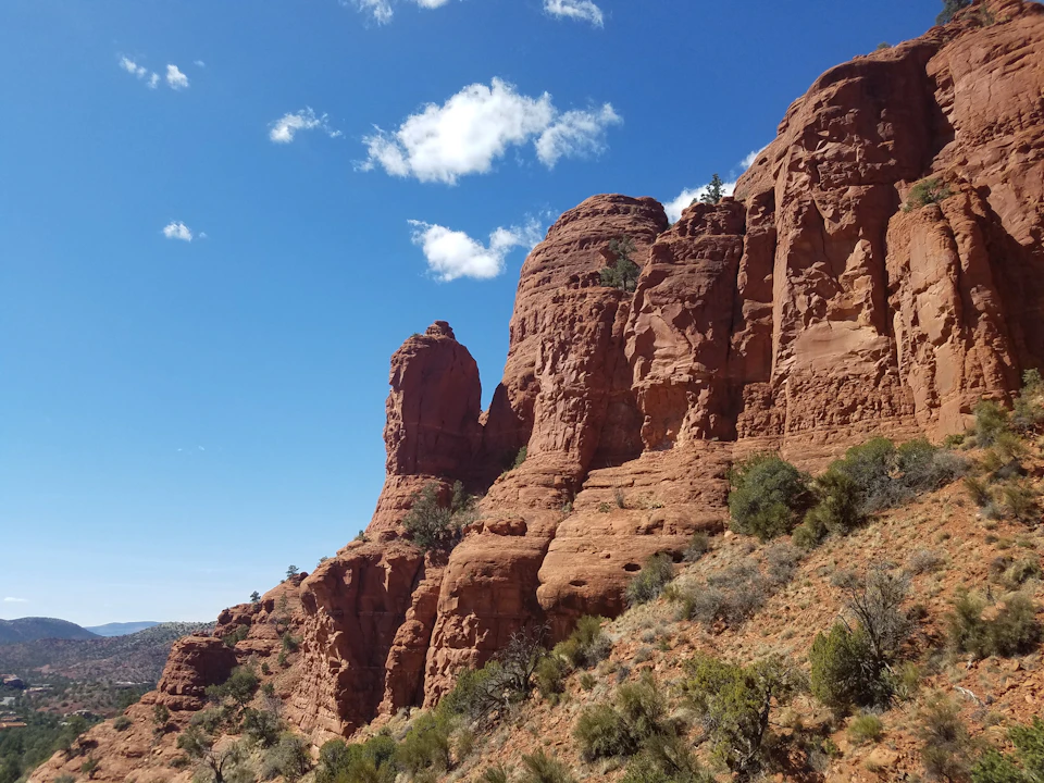 Red rock formations with sparse vegetation under a clear, blue sky with a few white clouds.