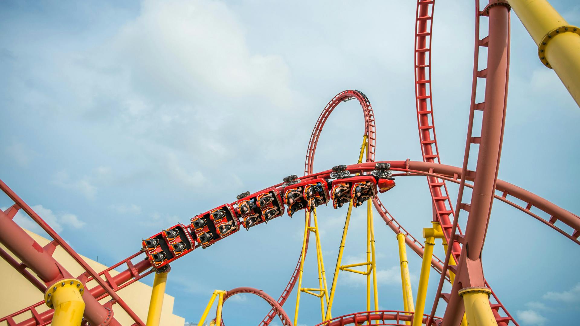 A red rollercoaster with riders loops through a yellow structure under a blue sky.