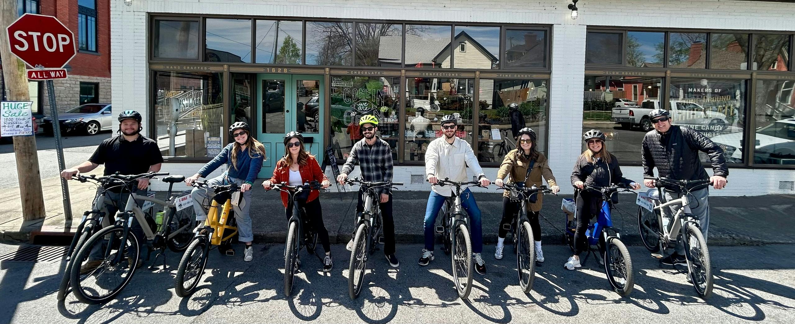 Five people wearing helmets are standing with bicycles in front of a building with large windows on a sunny day.