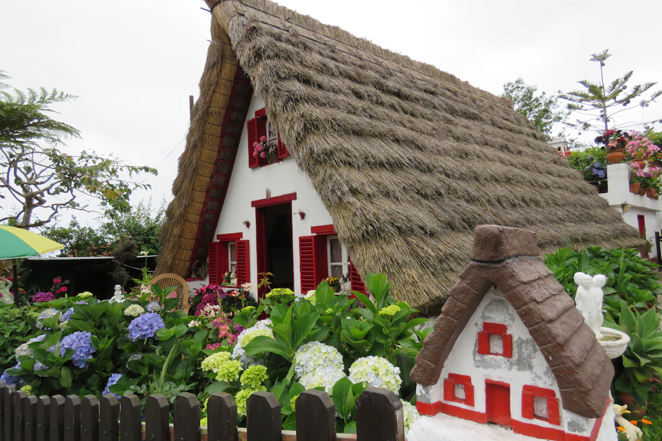 A thatched-roof cottage with red shutters and a small garden with colorful flowers. A small model house is visible in the foreground.