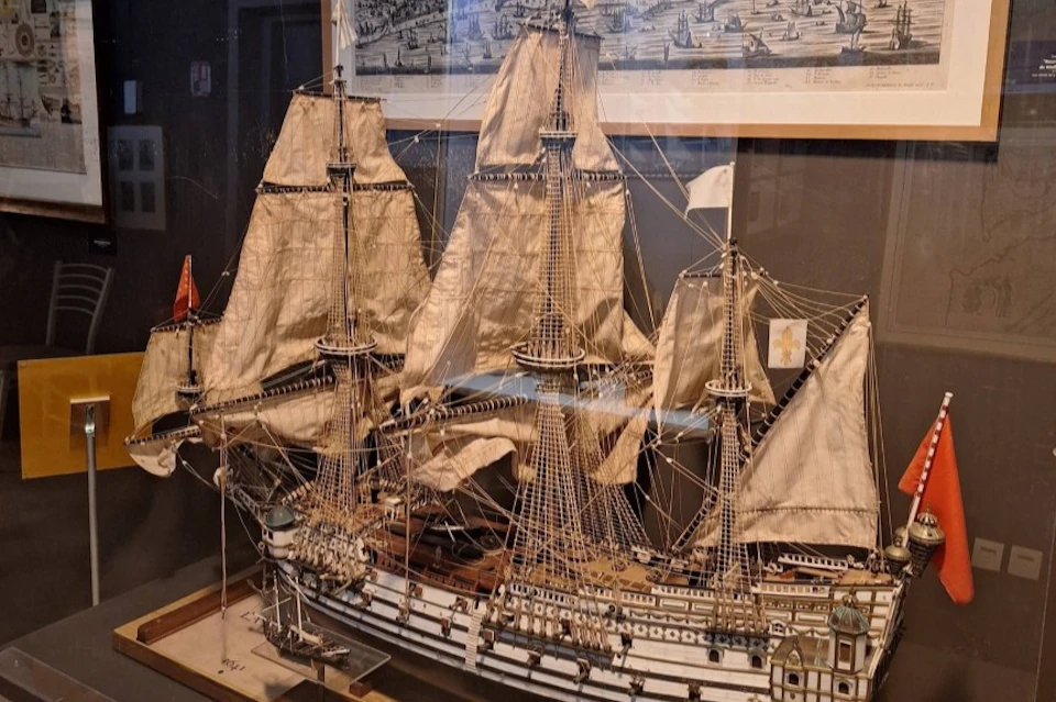 A detailed model of a sailing ship with three masts and beige sails, displayed in a museum setting with a historical map behind.