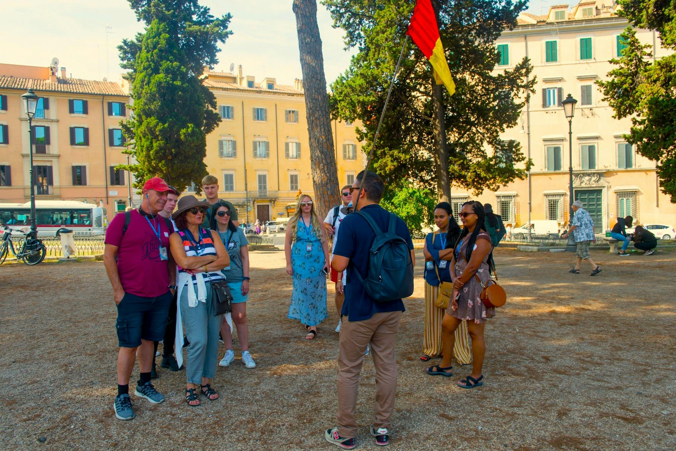 Un gruppo di persone all'aperto in una piazza con alberi ed edifici sullo sfondo, uno dei quali tiene in mano una bandiera rossa e gialla.