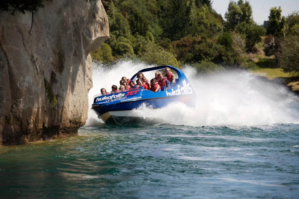 A blue jet boat with passengers speeding near a rocky cliff over a river, leaving a large white water spray behind.