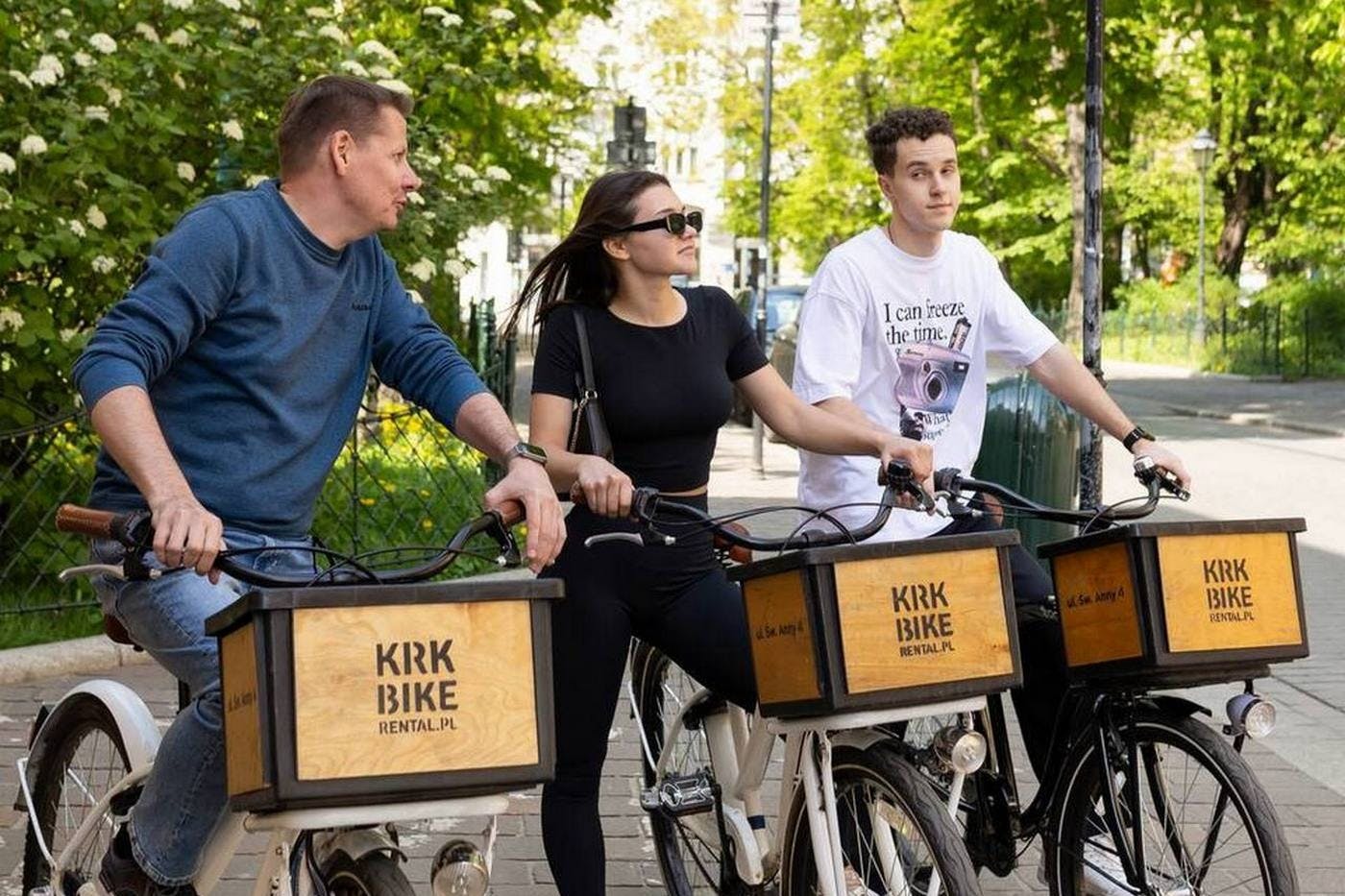Dva lidé jedoucí na půjčeném kole v blízkosti zeleného parku. Na kolech jsou dřevěné krabice s nápisem „Kirk Bike Rental“.