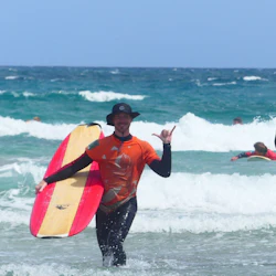 Person in orange shirt and black hat holding surfboard, making a gesture, emerging from ocean waves. Other surfers in the background.