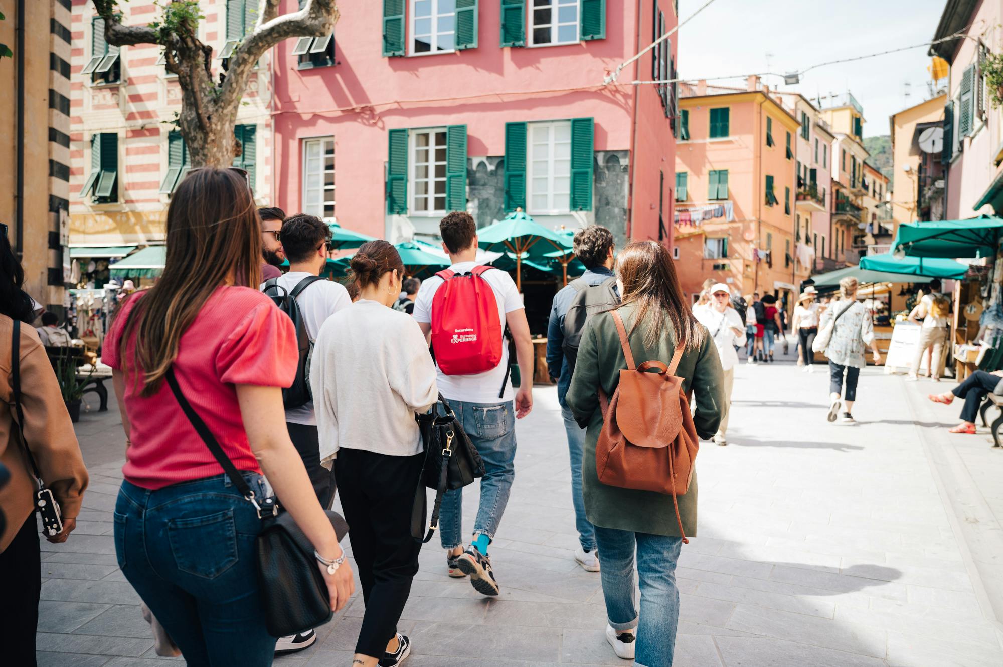 一群人走过一个阳光明媚、色彩缤纷的广场，广场上有粉色和橙色的建筑，绿色的百叶窗。