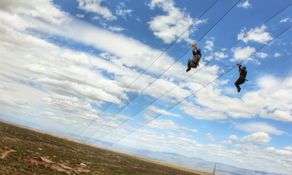 Person zip-lining across a vast landscape under a partly cloudy sky with mountains in the distance.