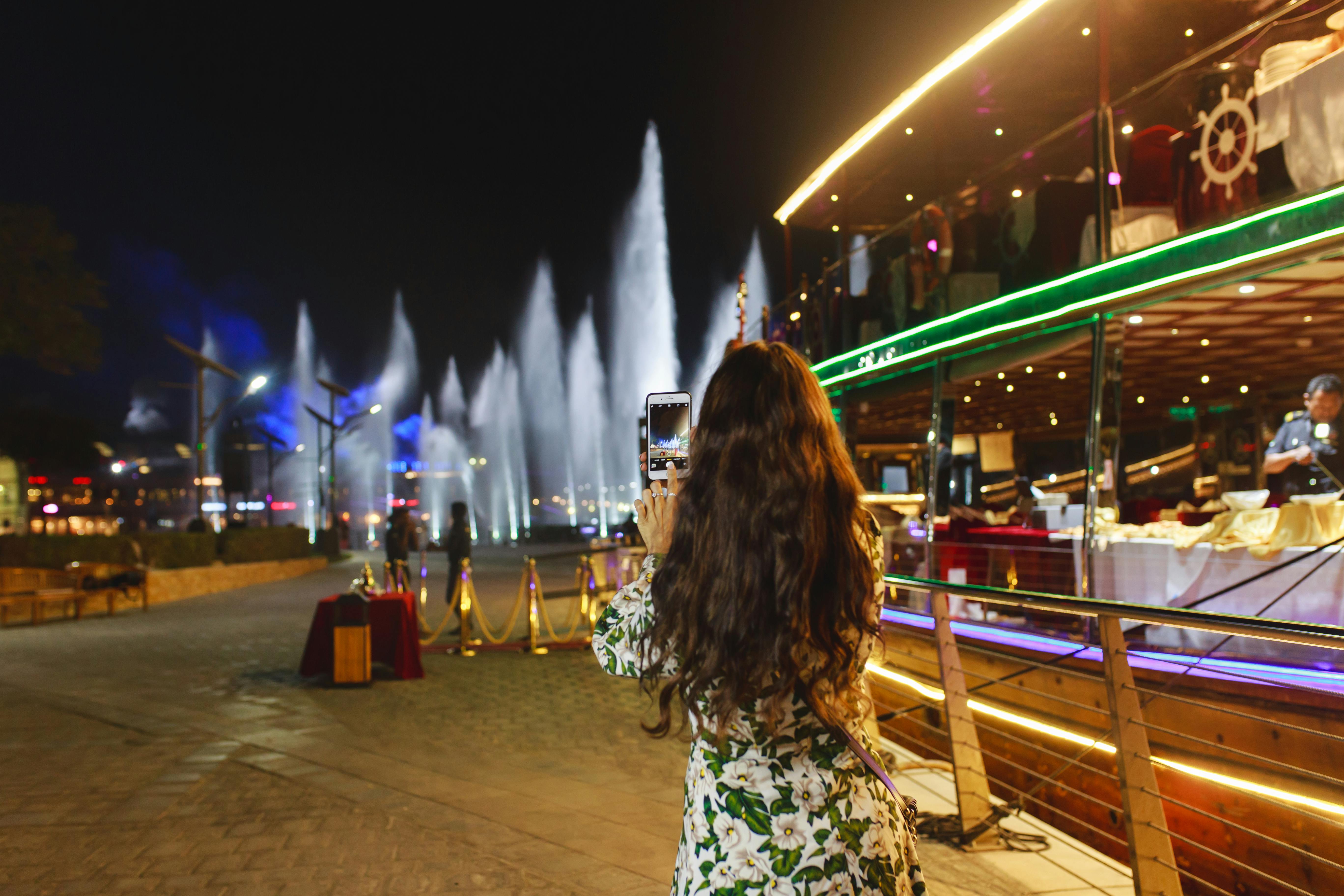 A woman with long hair takes a photo of lit water fountains at night, near a multilevel structure adorned with lights.