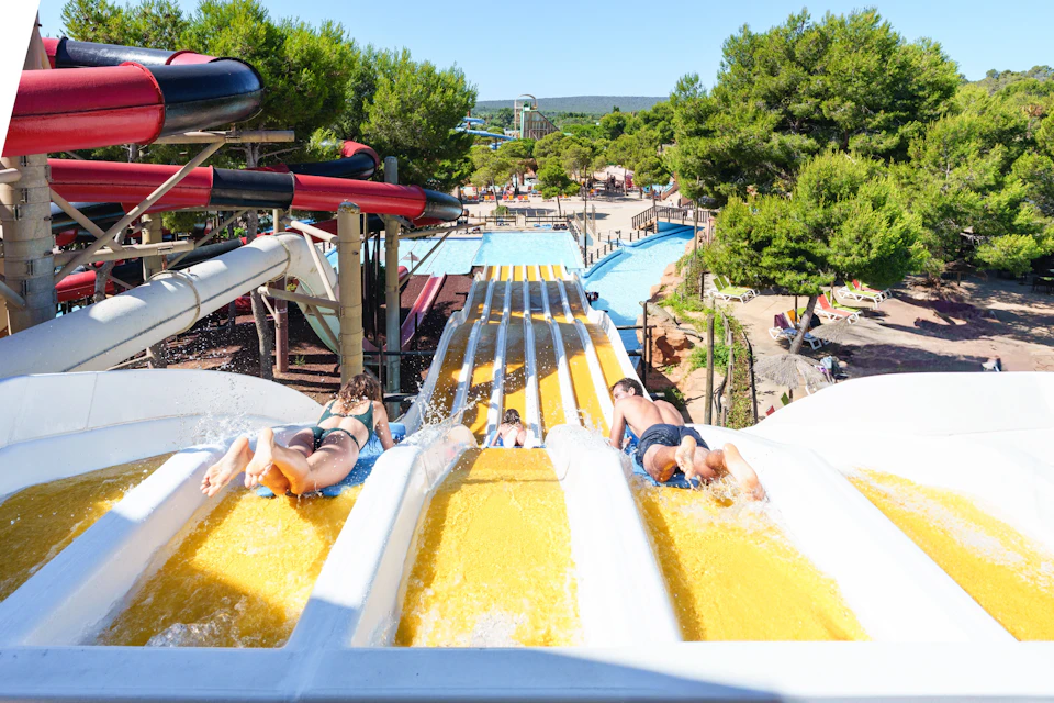 People sliding down multi-lane yellow water slides at a water park with trees and pools in the background.