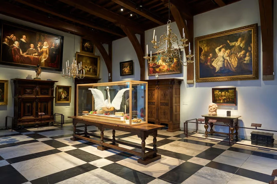 A museum gallery with paintings on the walls, chandeliers, and a display of a swan in a glass case on a wooden table.