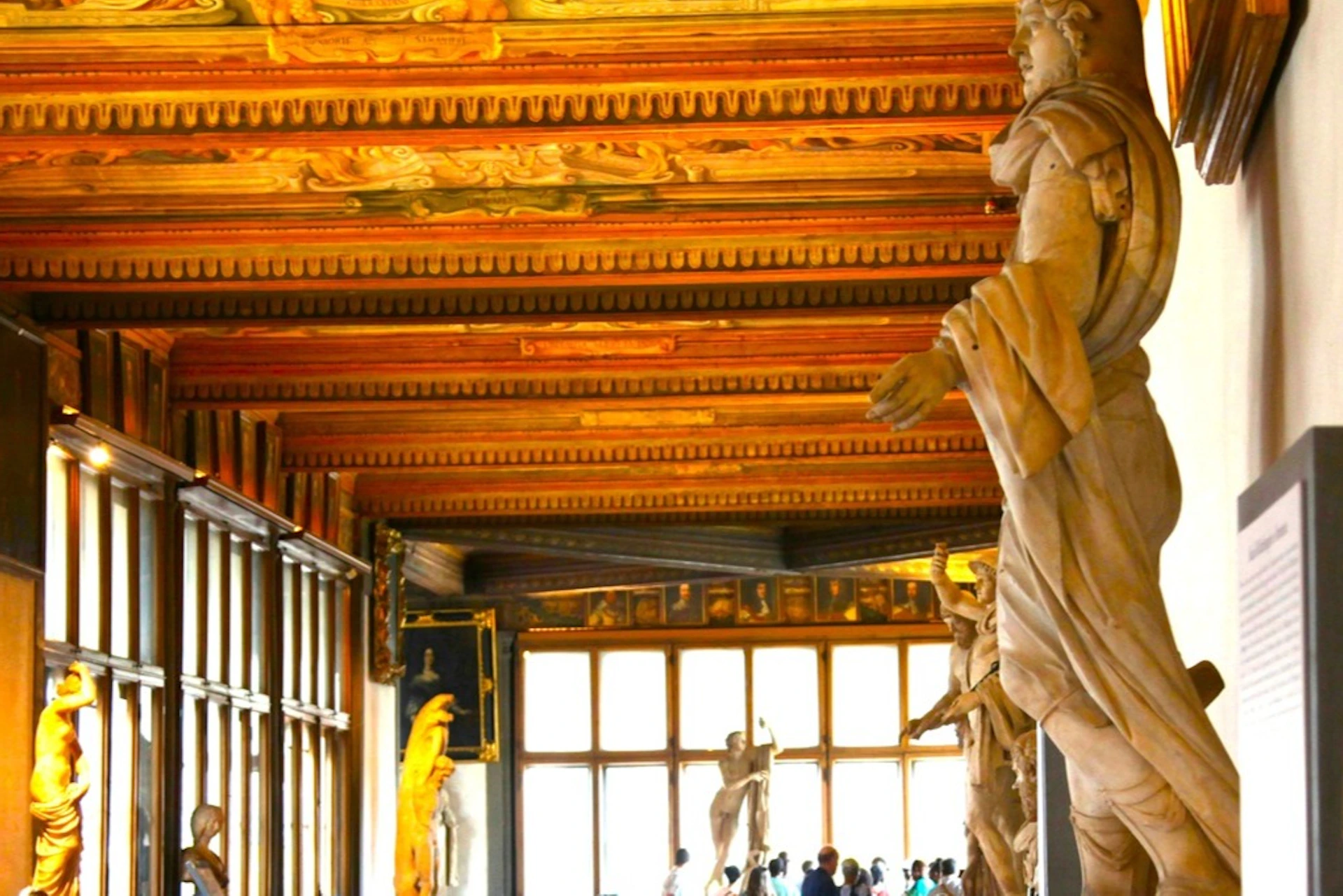 A museum gallery with statues, ornate ceilings, and visitors walking and observing exhibits.