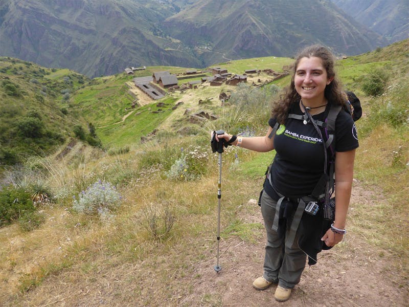 A woman with a backpack and hiking pole smiles on a grassy hilltop with ancient ruins and mountains in the background.