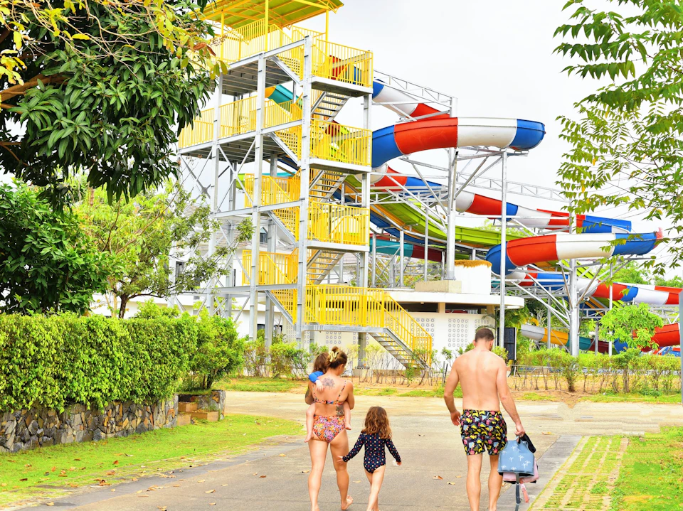A family walking near a colorful water slide structure in a park-like setting with trees and greenery.