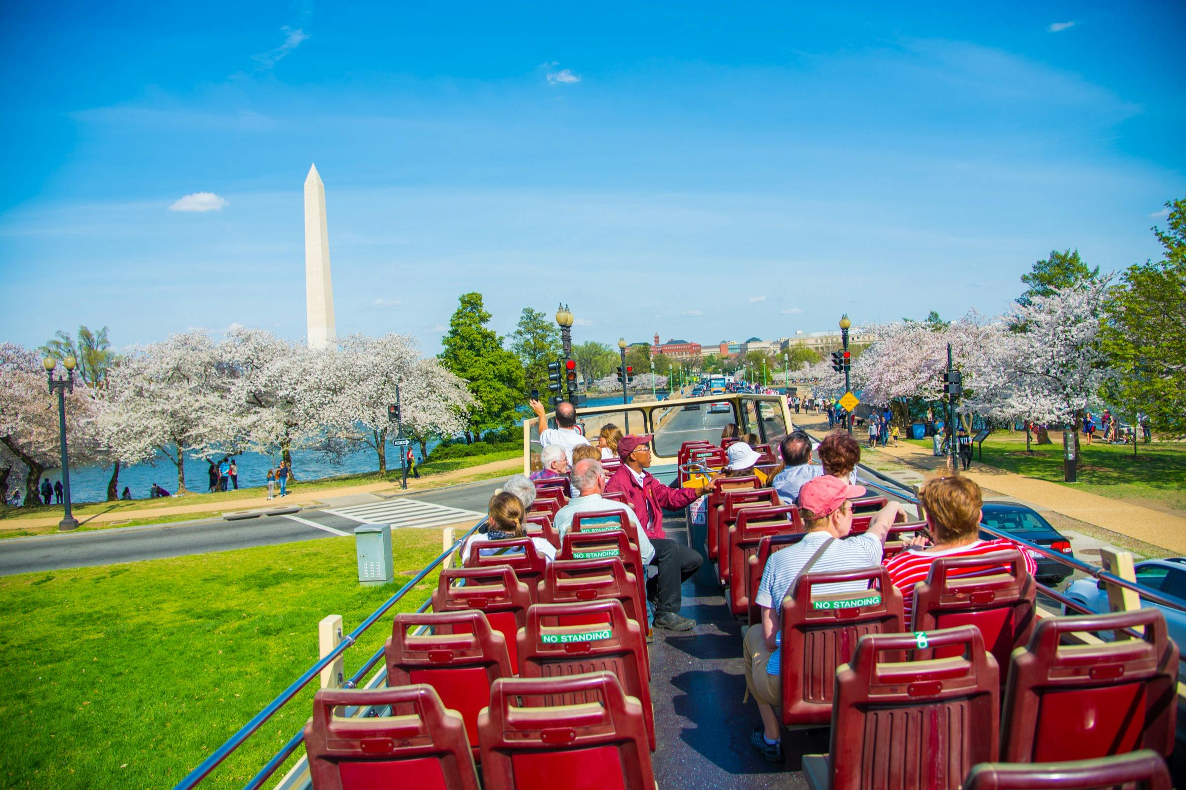 Tourists on an open-top bus viewing the Washington Monument and blooming cherry trees under a clear blue sky.