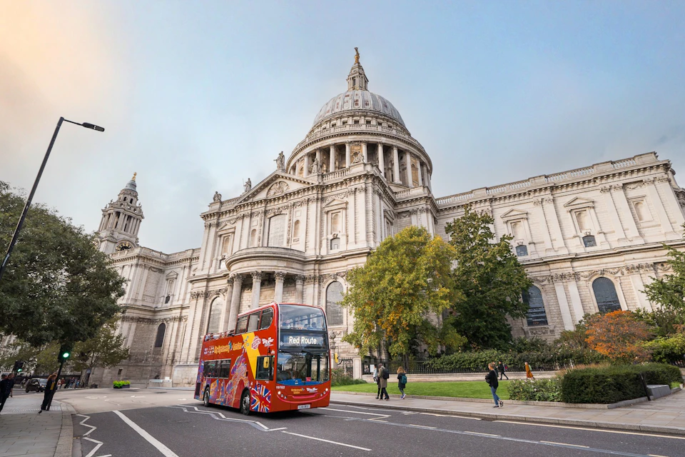 A red double-decker bus in front of St. Paul's Cathedral in London, with a few pedestrians walking nearby and trees in view.