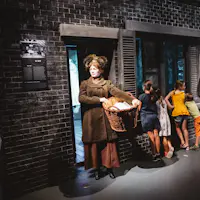 A woman in historical attire holds a basket outside a brick building. Children look into a window beside her.