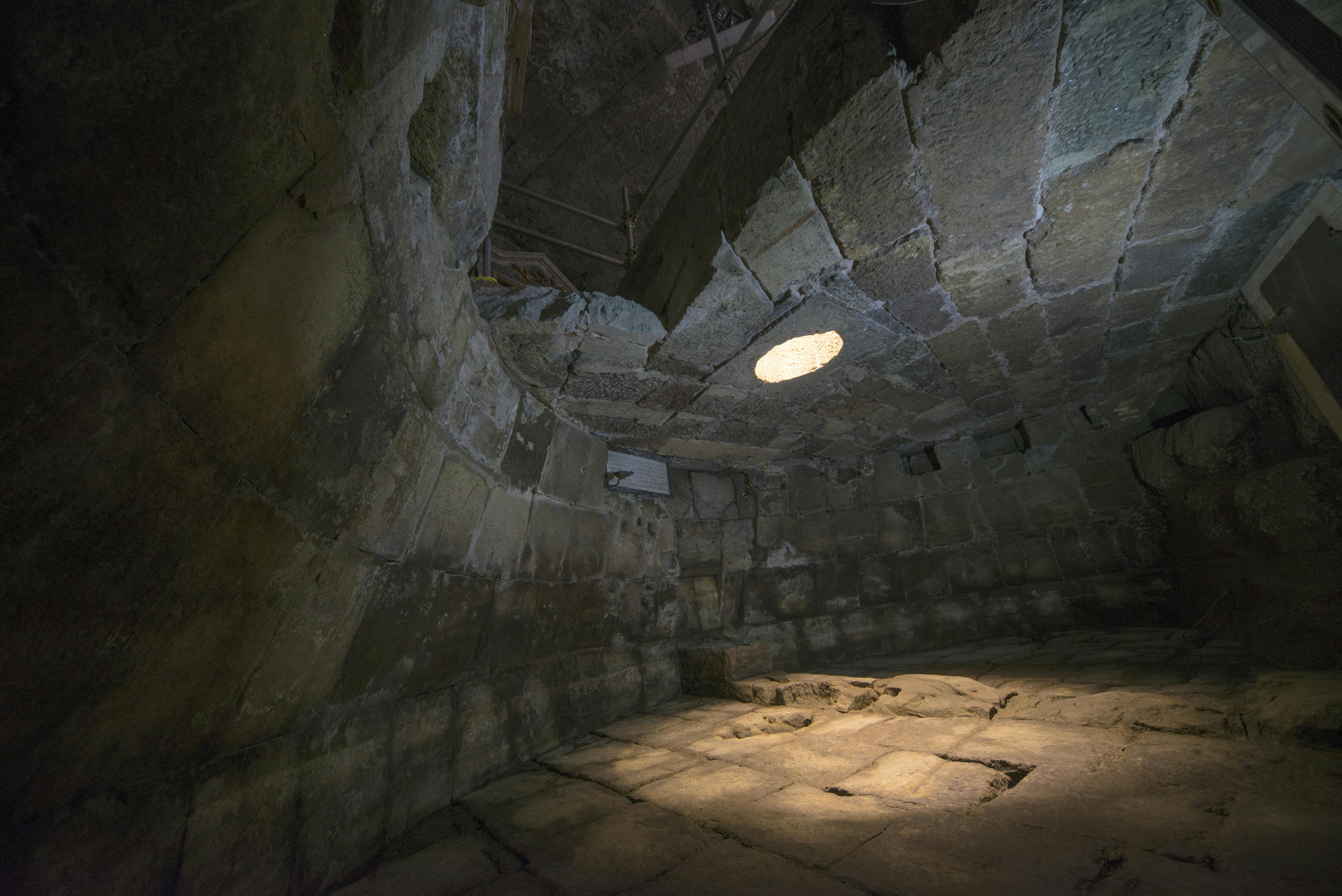 A dimly lit, ancient stone chamber with a circular hole in the ceiling allowing light to enter.