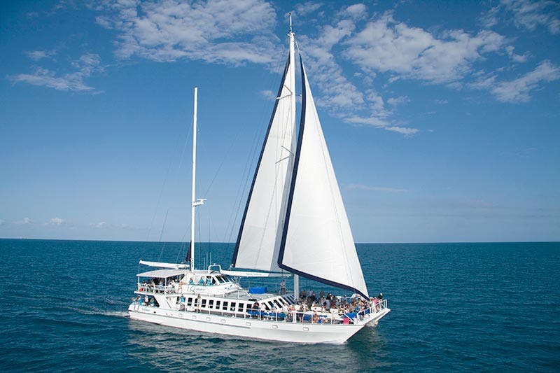 A white sailboat with open sails and several people onboard is sailing on a calm blue ocean under a partly cloudy sky.