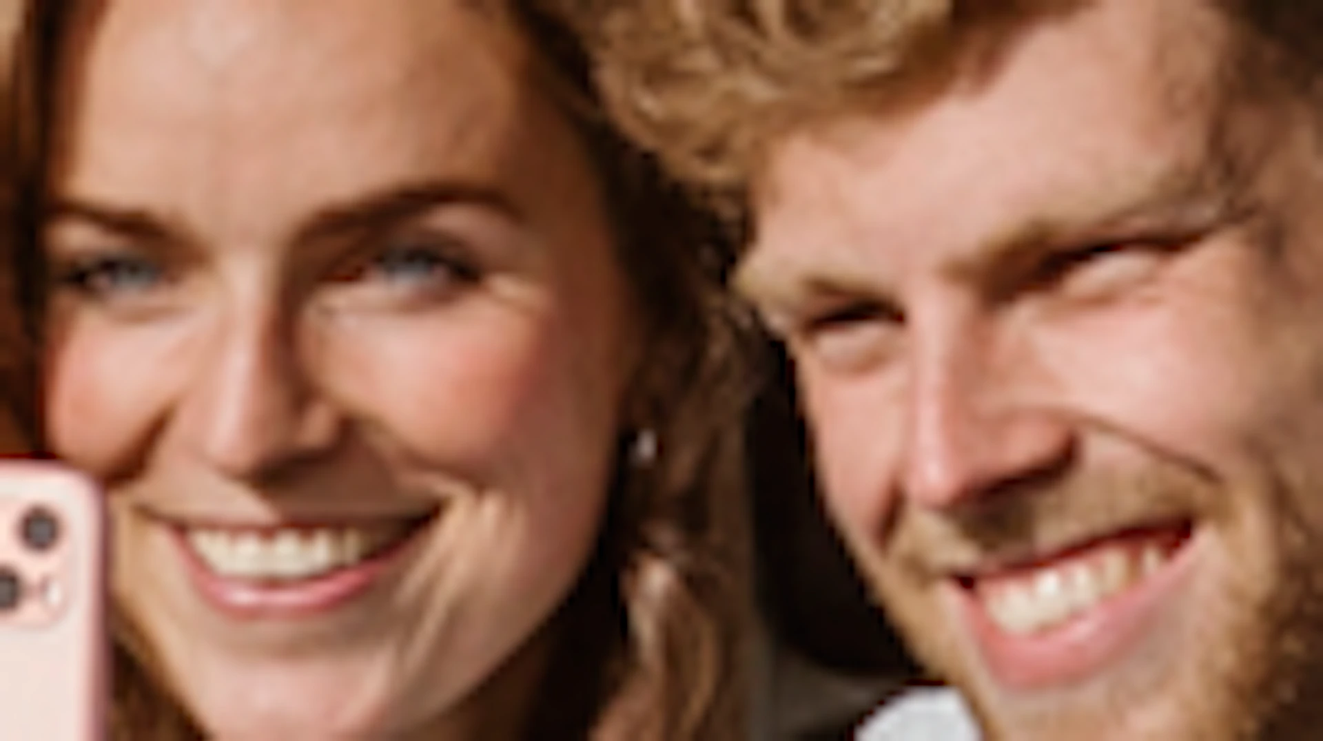 A couple takes a selfie while sitting in a camo-painted car with open doors amid a vibrant flower field.