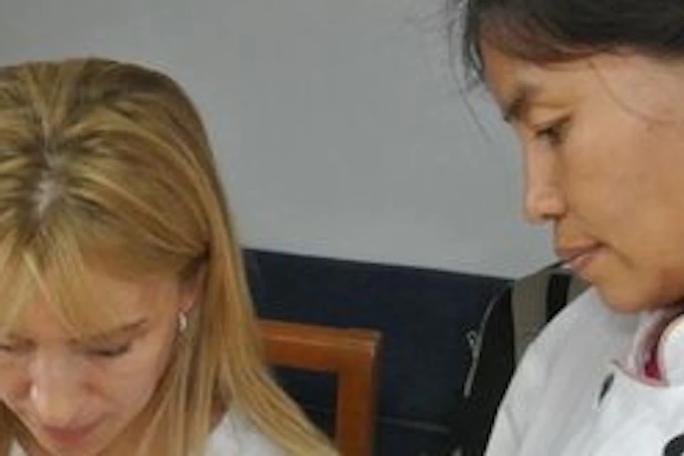 A woman in a chef coat instructs another woman on carving carrots on a cutting board.