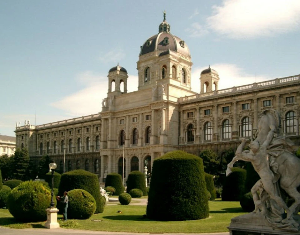 Elegant historic building with a central dome, surrounded by neatly trimmed hedges, statues, and a manicured garden. Clear skies above.