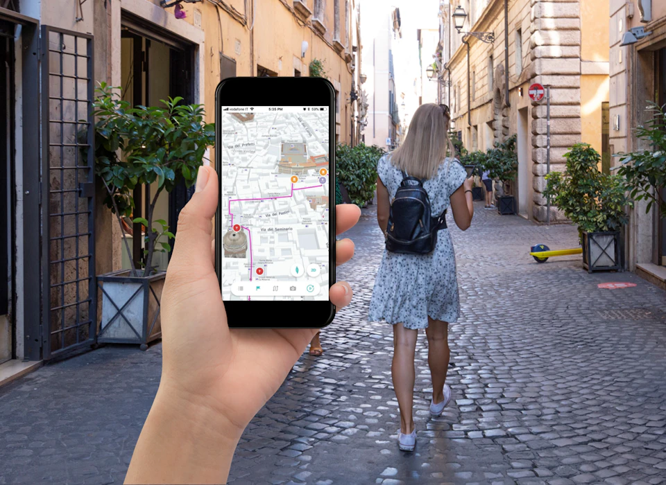 A person holds a smartphone showing a map app while walking behind a woman in a blue dress and black backpack on a narrow street.