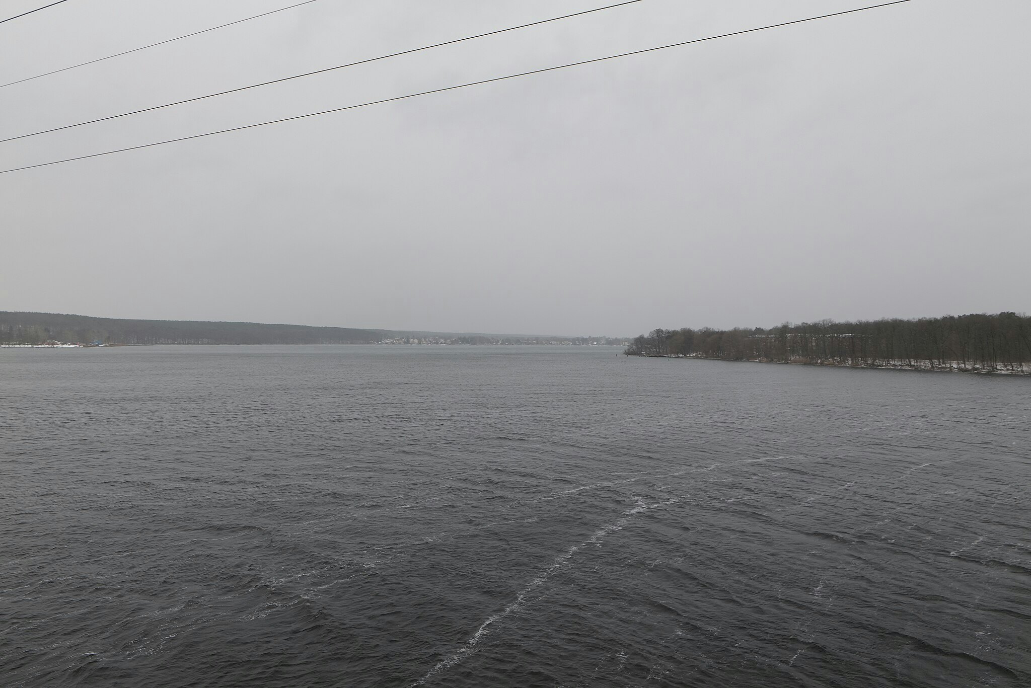 Plan d'eau calme avec un ciel couvert, un rivage lointain avec des arbres et des lignes électriques aériennes.