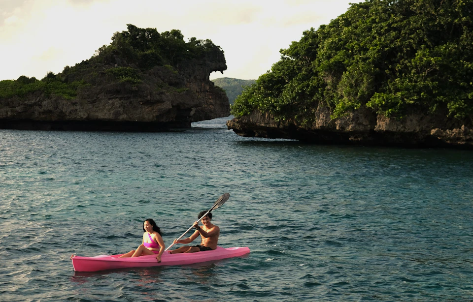Two people kayaking in a pink kayak on clear water near rocky, tree-covered islands under a partly cloudy sky.
