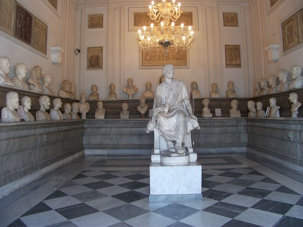 A marble statue of a seated figure is surrounded by numerous busts on display in an ornate room with a chandelier and checkered floor.