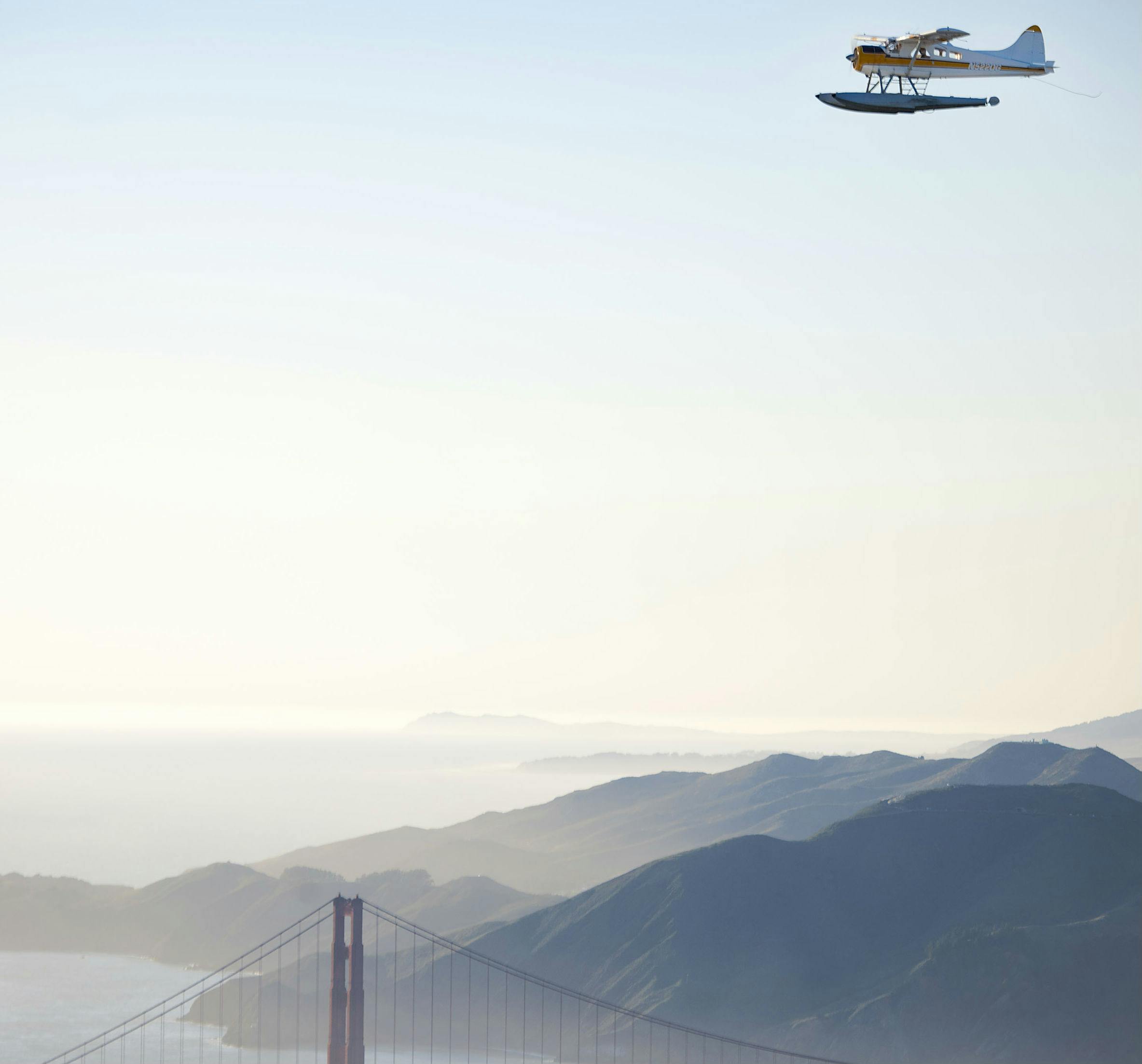 A seaplane flying over a coastal landscape with a bridge and mountainous terrain in the background.