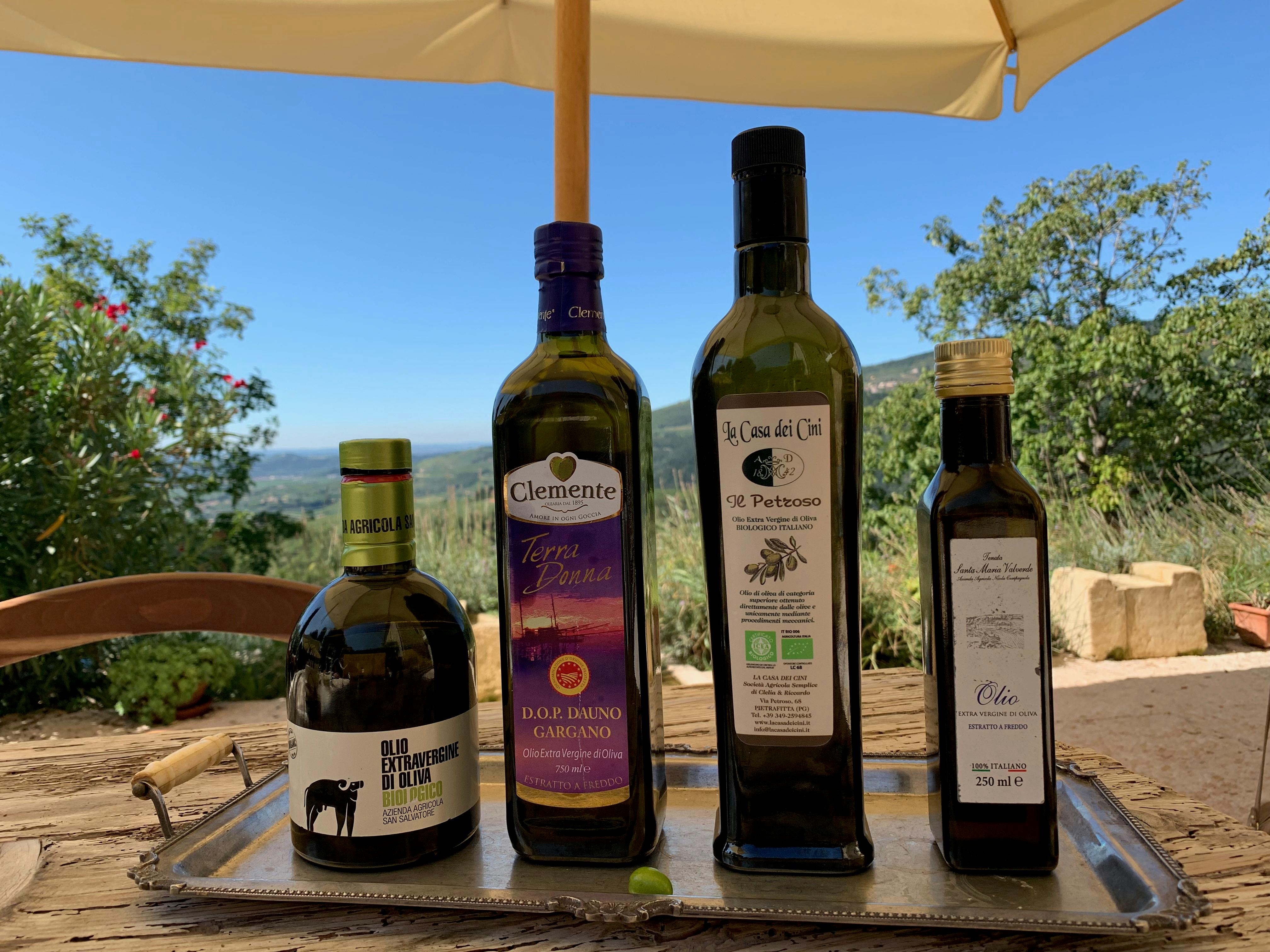 Four bottles of olive oil on a tray outdoors, with a scenic background of trees and a clear sky under a patio umbrella.