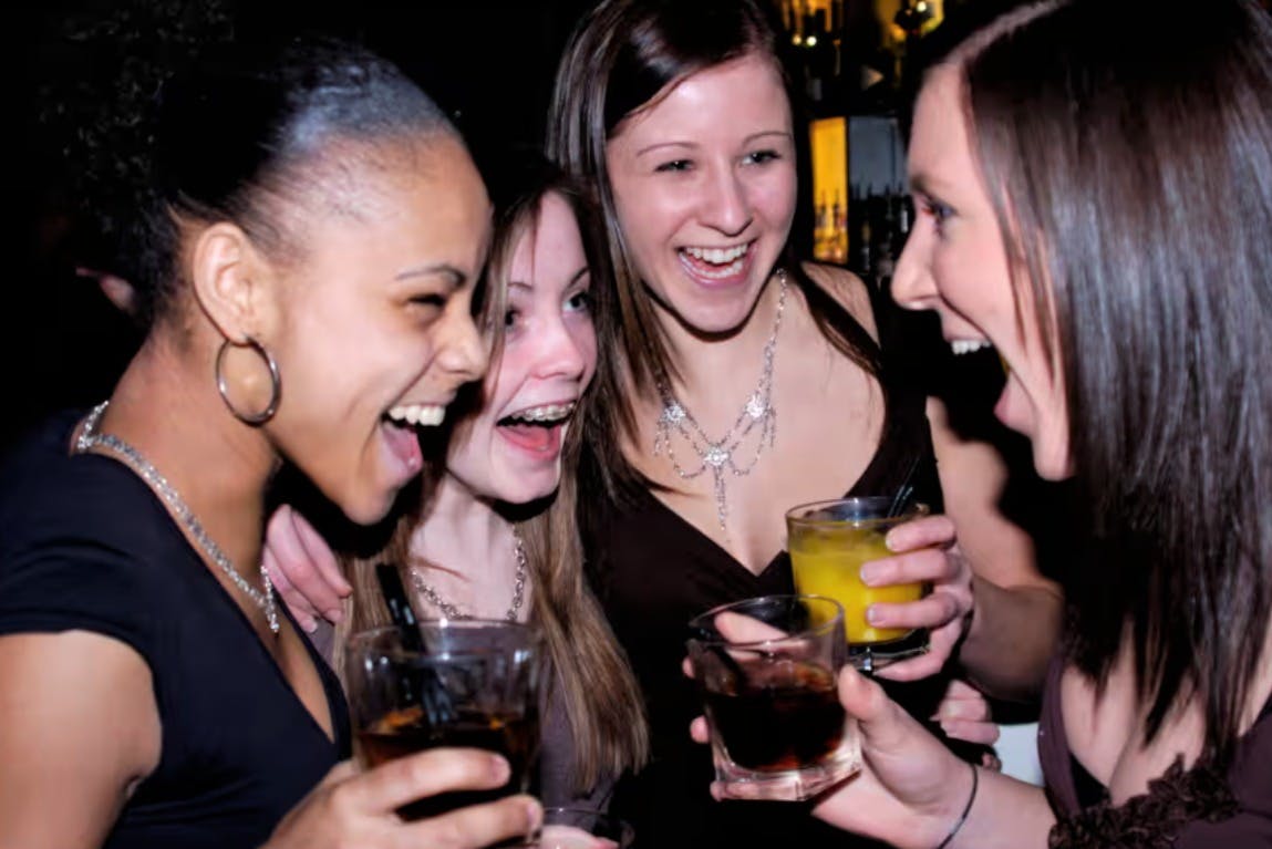 Four women laughing and holding drinks at a bar.