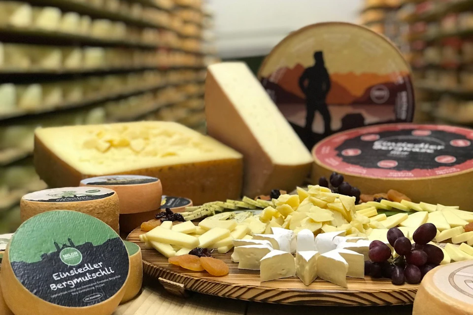 A platter of assorted cheeses with fruits and a background of cheese wheels in a store.