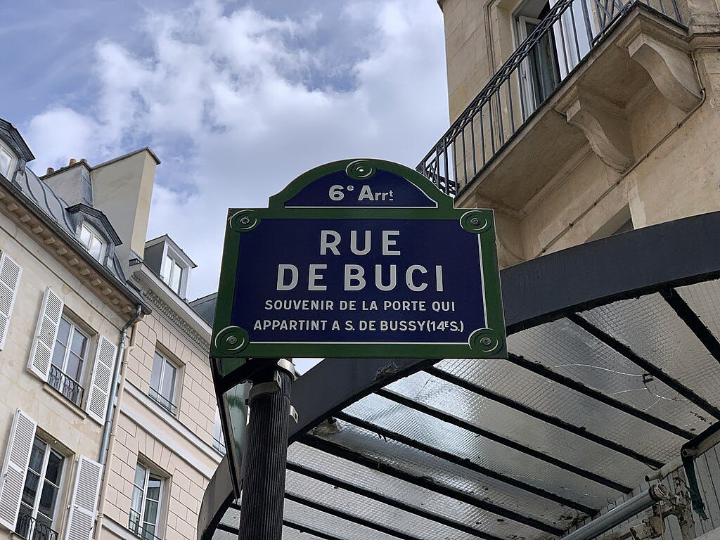 Street sign reading "Rue de Buci" in Paris, 6th arrondissement, with buildings and a metal structure in the background.