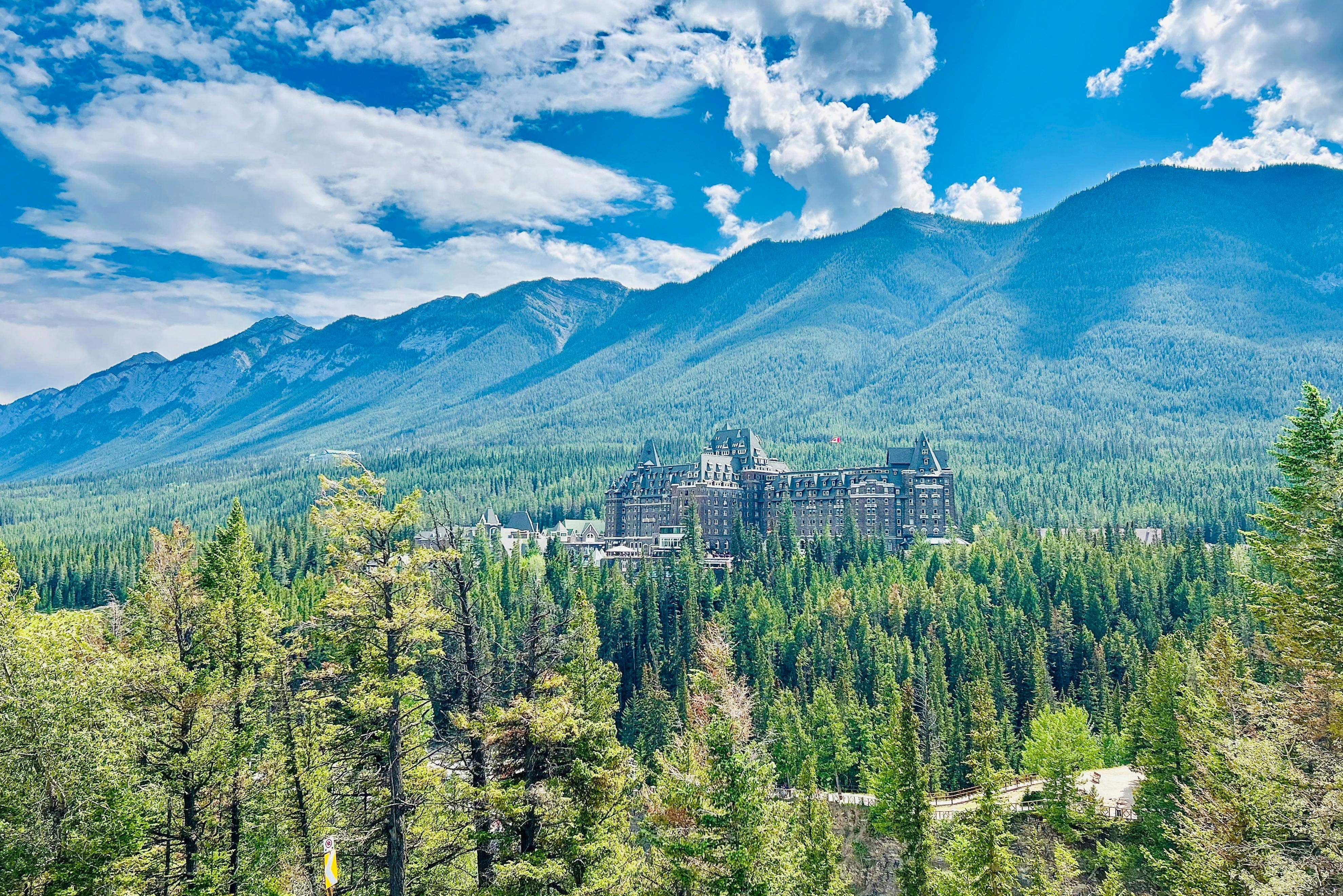 A large castle-like building surrounded by dense pine trees with mountains and a blue sky with clouds in the background.