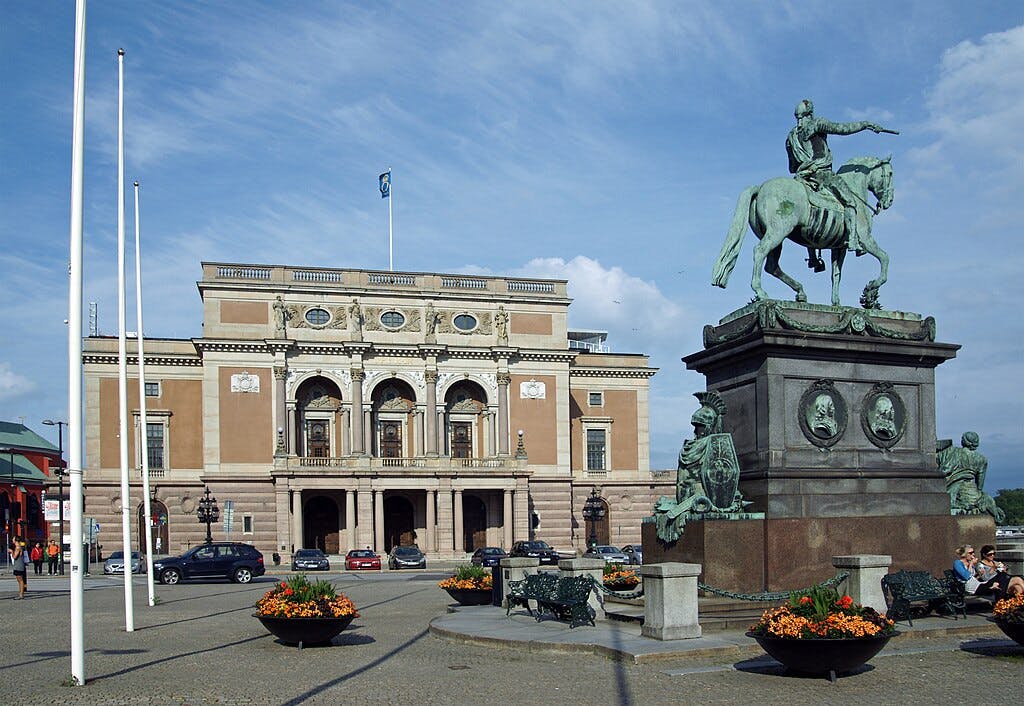A grand building with arched windows and a flag on top, in front of a statue featuring a man on horseback, and potted flowers.