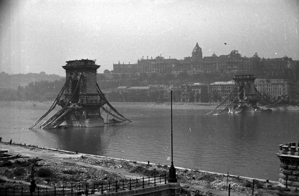 Ruined bridge over a river with large buildings on the opposite bank. Debris and construction materials in the foreground.
