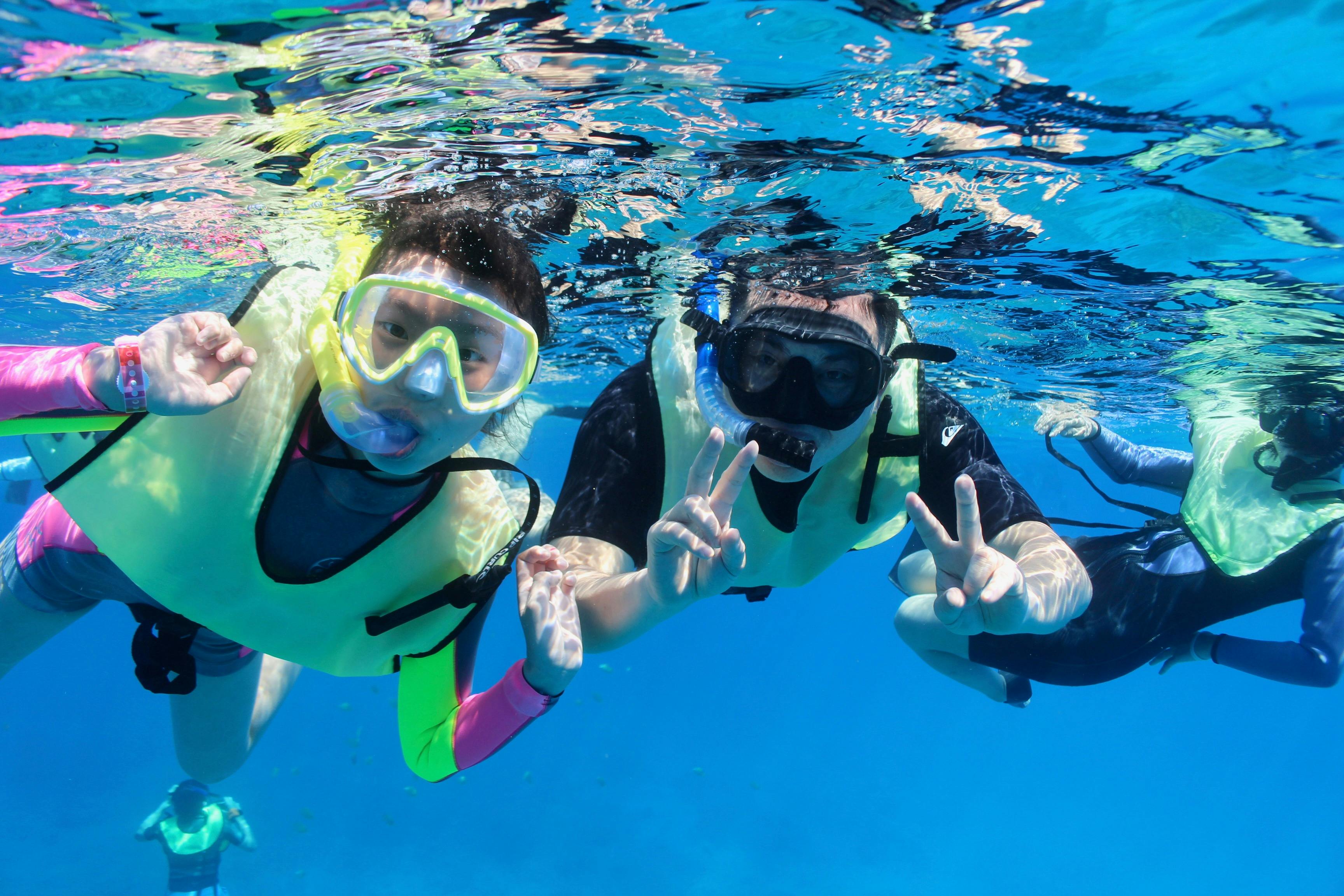 Dva lidé, kteří mají na sobě šnorchlovací výstroj a záchranné vesty, dělají mírové značky směrem ke kameře v průzračné modré vodě.