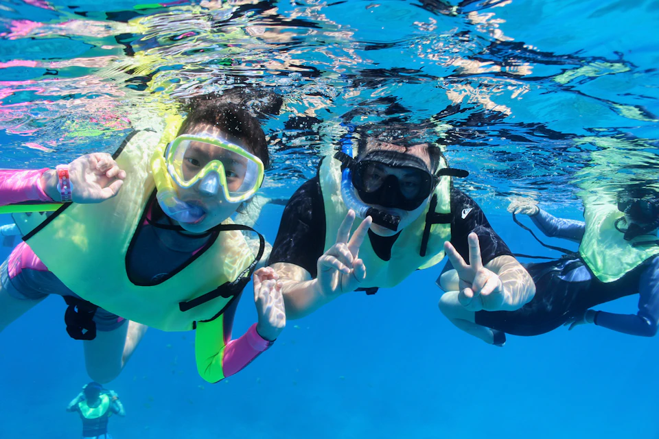 Two people wearing snorkeling gear and life vests underwater, making peace signs towards the camera in clear blue water.