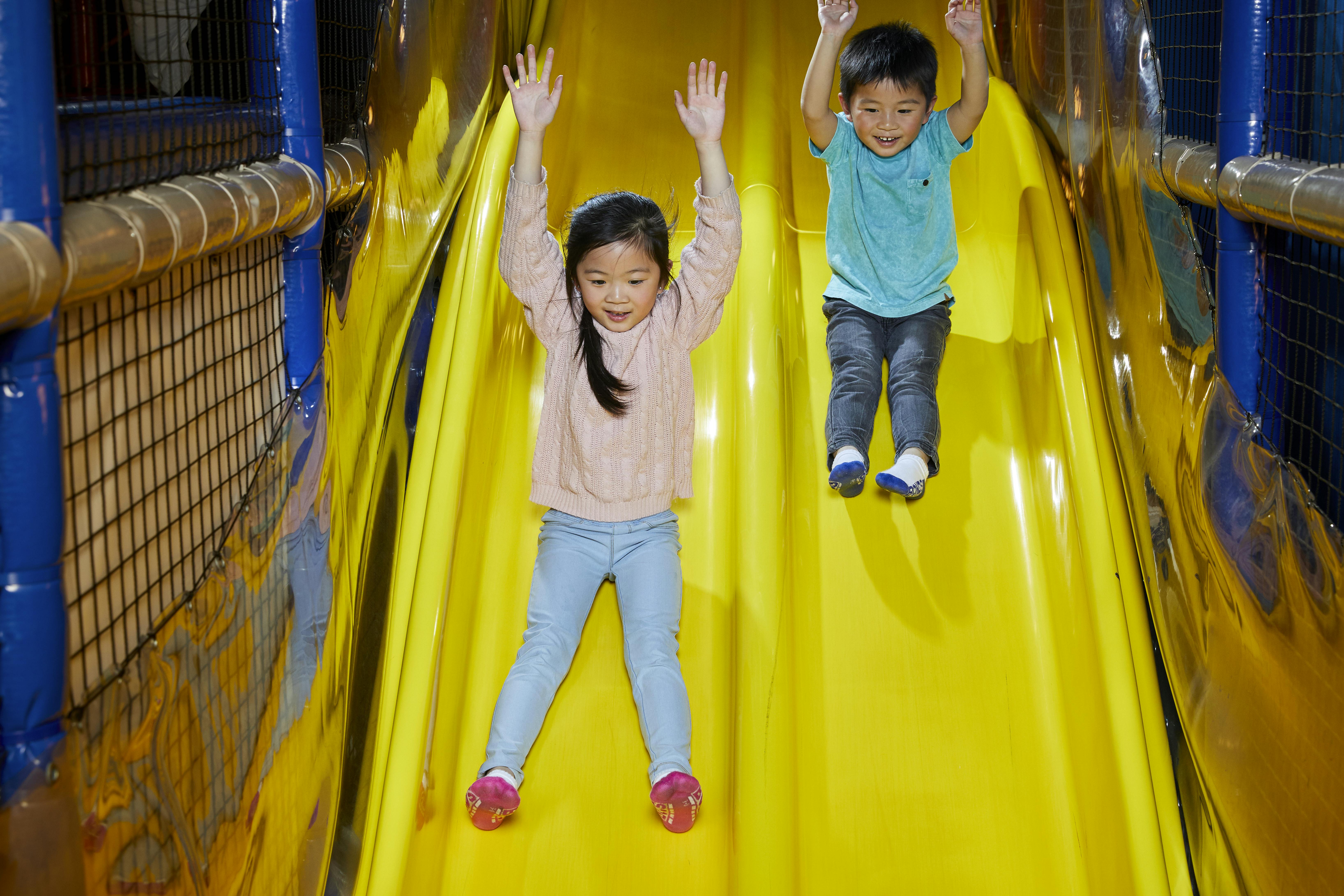 Two children sliding down a bright yellow slide, both with their arms raised and expressions of excitement on their faces.