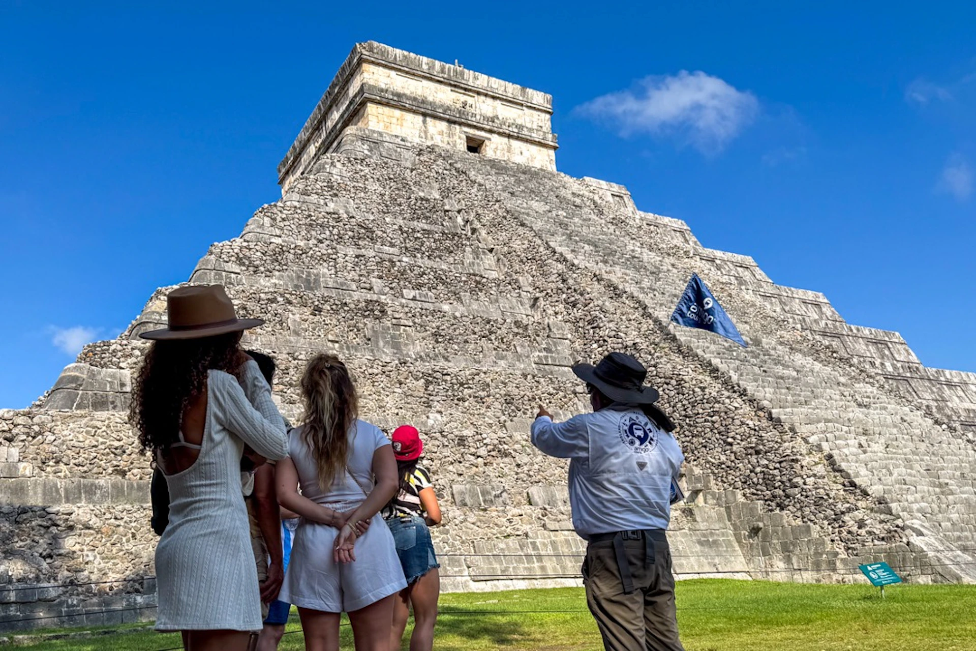 A group of people listen to a guide in front of a large, ancient stone pyramid under a clear blue sky.
