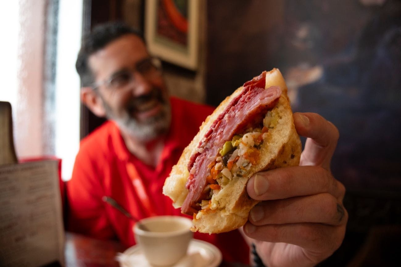 Un uomo in camicia rossa tiene in mano un panino pieno di carne e verdure, con una tazza di caffè sul tavolo di fronte a lui.