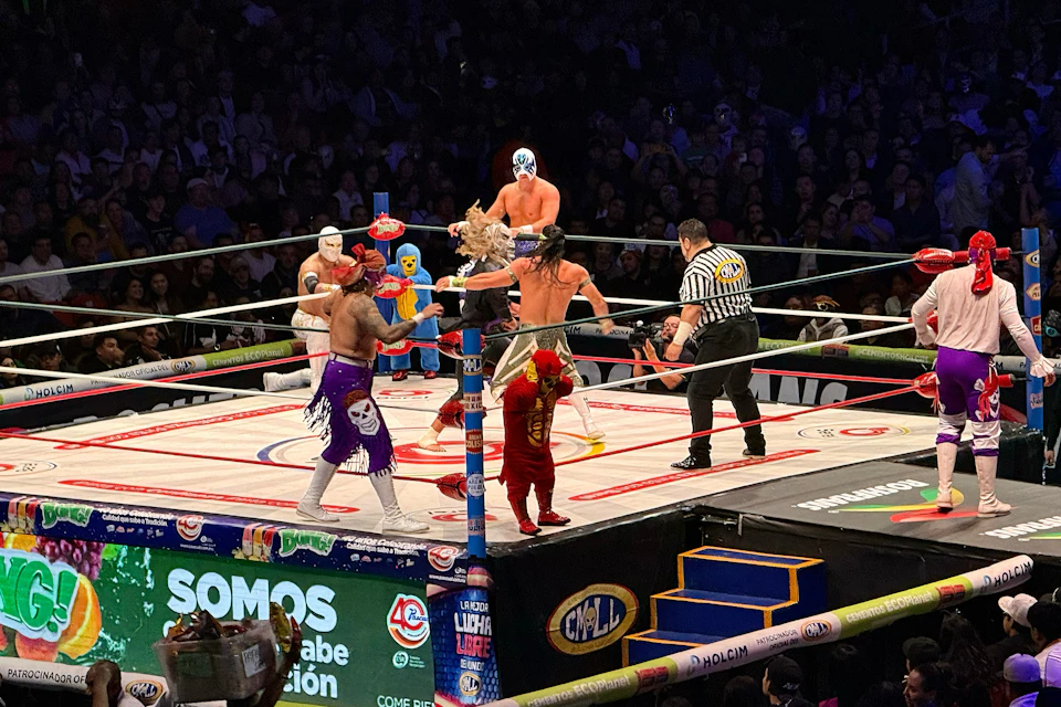 A wrestling match with multiple masked wrestlers and a referee in a ring, surrounded by a crowd in an arena.