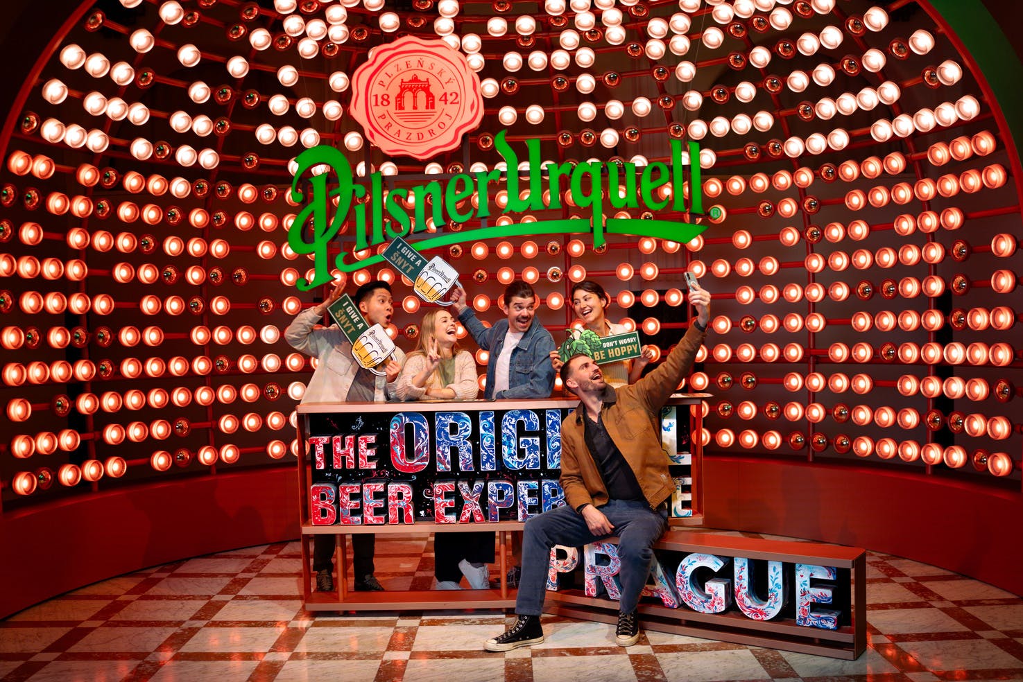 一群人在明亮的空间里欢快地摆姿势，那里有Pilsner Urquell标牌和装饰灯。